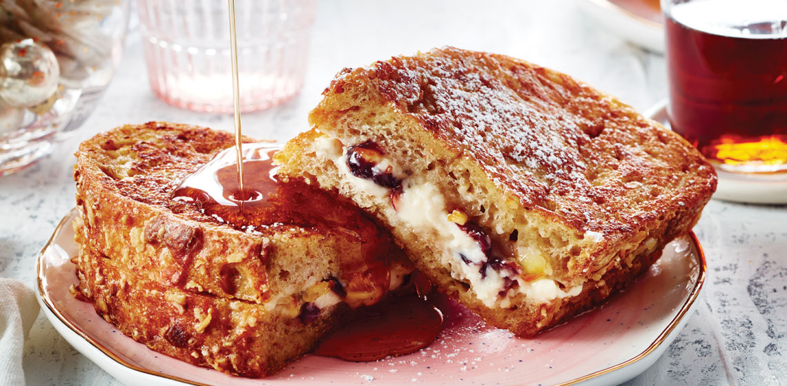 A close-up of a beige plate topped with a cream cheese and dried fruit–stuffed French toast with a drizzle of maple syrup falling on one side of the plate.