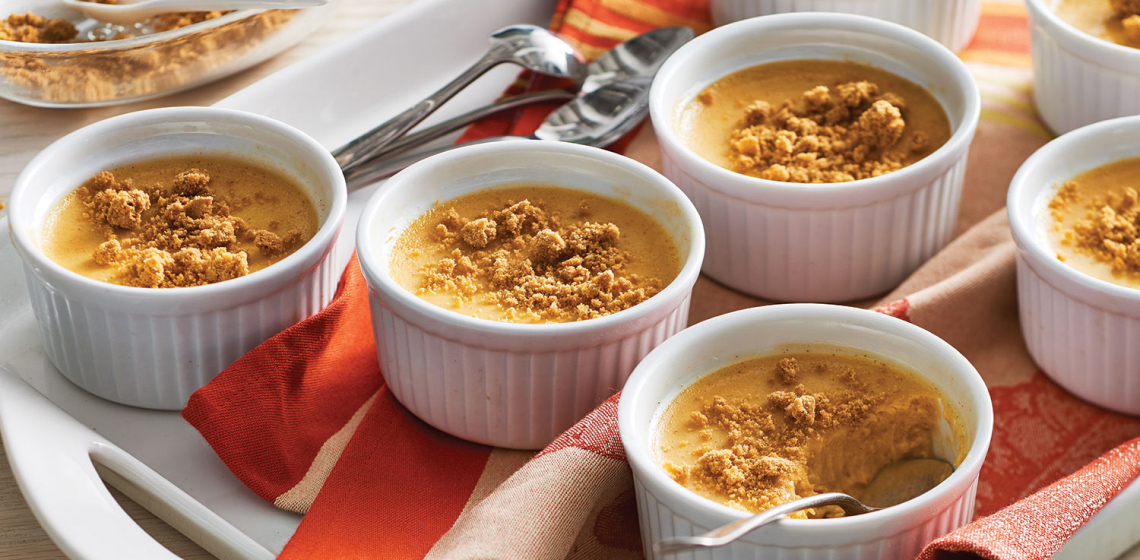 White ramekins of sweet potato custard topped with gingersnap crumble on top of a red and white kitchen towel next to a small pile of spoons.