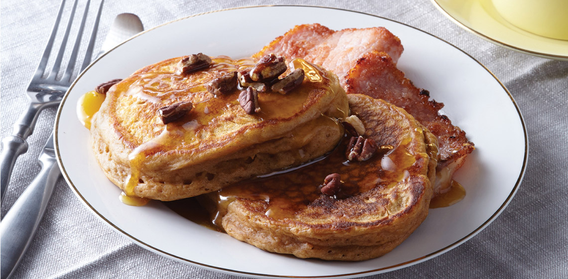 Two sweet potato gingerbread pancakes on a white plate topped with maple syrup and pecans next to two peameal bacon slices. 