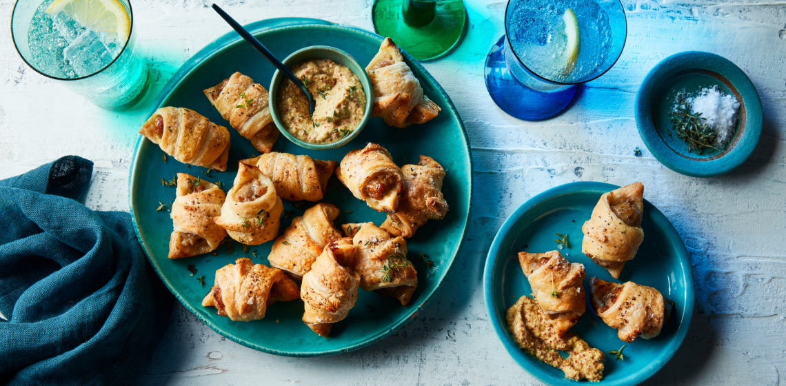 Tablescape with sparkling waters, napkin and round aqua green-blue serving plate with bratwurst pigs in blankets and a small bowl of grainy Dijon mustard for serving