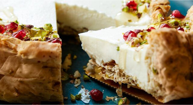 Sound blue serving platter on white marble surface with baklava cheesecake slice coming out of whole cake, topped with pomegranate arils and chopped pistachios