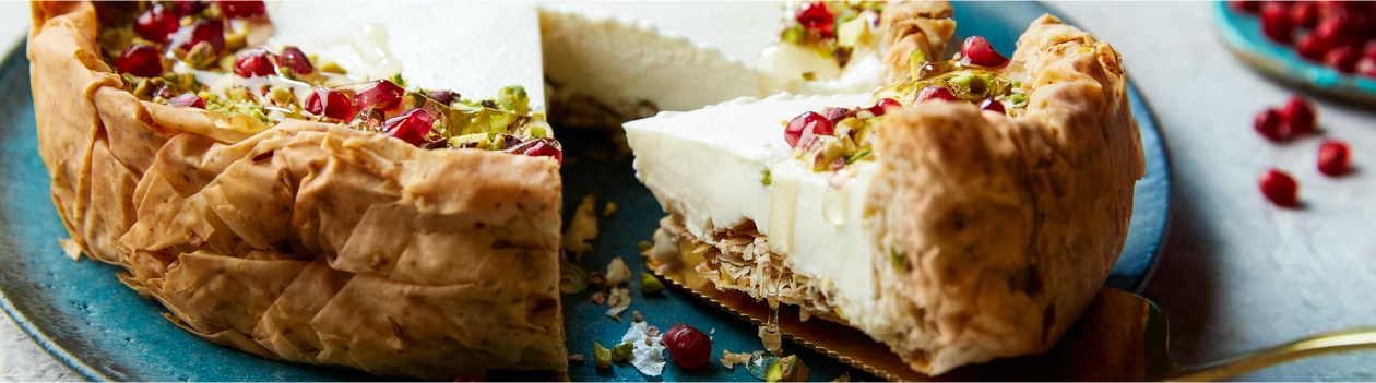 Sound blue serving platter on white marble surface with baklava cheesecake slice coming out of whole cake, topped with pomegranate arils and chopped pistachios