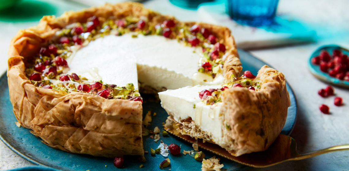 Sound blue serving platter on white marble surface with baklava cheesecake slice coming out of whole cake, topped with pomegranate arils and chopped pistachios