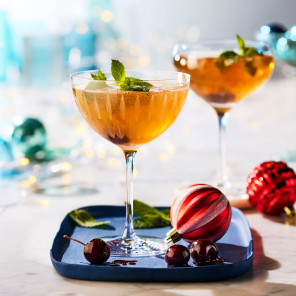 Easy holiday cocktails in two coupe glasses filled with a light amber cocktail and a mint leaf garnish on a marble surface with scattered holiday decorations.