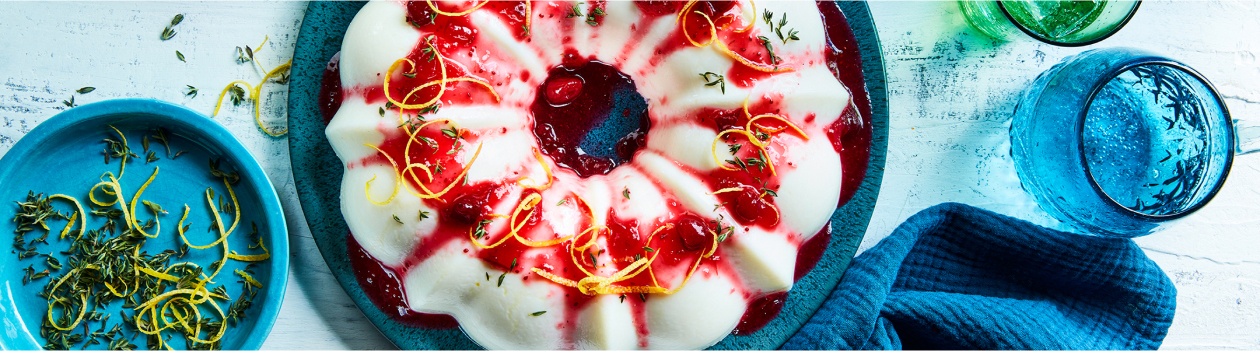 Round blue cake platter with chilled cranberry-lemon blancmange, with cranberry sauce, lemon peel and thyme leaf garnish.