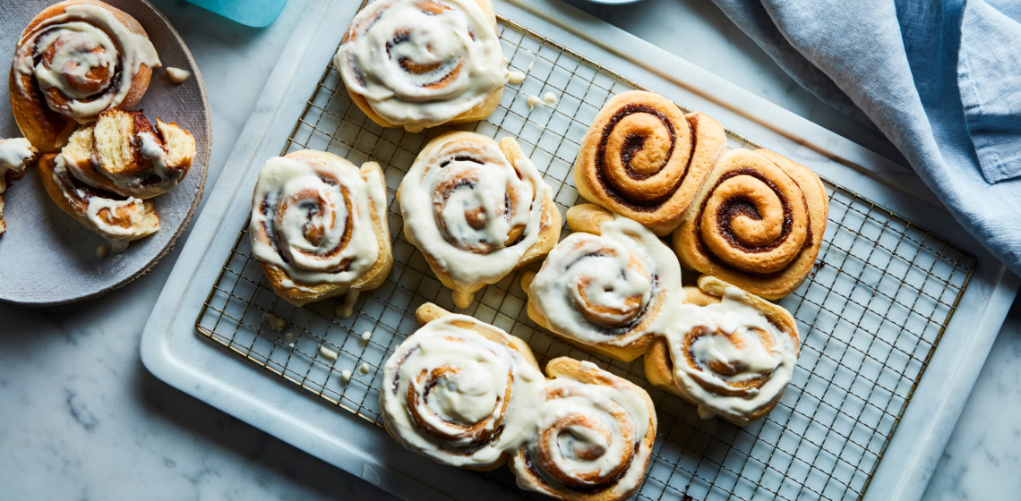 Marble surface with cooling rack of glazed cinnamon rolls, with side plate of half-eaten roll to the side