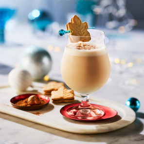 An easy milky holiday cocktail in a bell-shaped short-stemmed glass with a maple crème cookie on a skewer across the rim, all set on a white marble surface with holiday decorations.