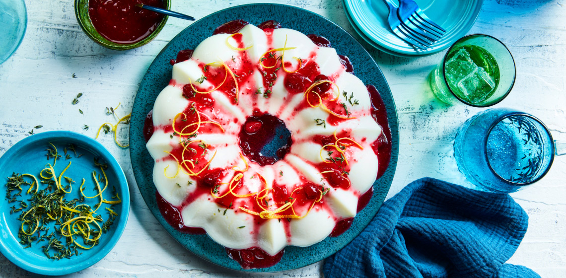 Round blue cake platter with chilled cranberry-lemon blancmange, with cranberry sauce, lemon peel and thyme leaf garnish.