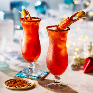 Two tall bell-shaped glasses with a dirty Caesar cocktail and king crab leg garnish on a white marble surface with holiday decorations in the background.
