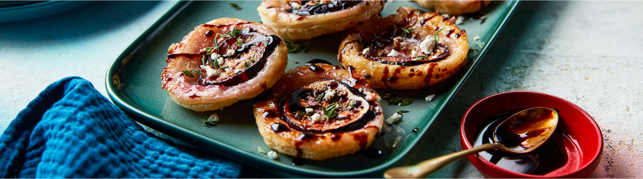 bite-sized rounds of baked puff pastry with fig, feta and balsamic glaze on a rectangular aqua serving platter with a blue napkin to the side