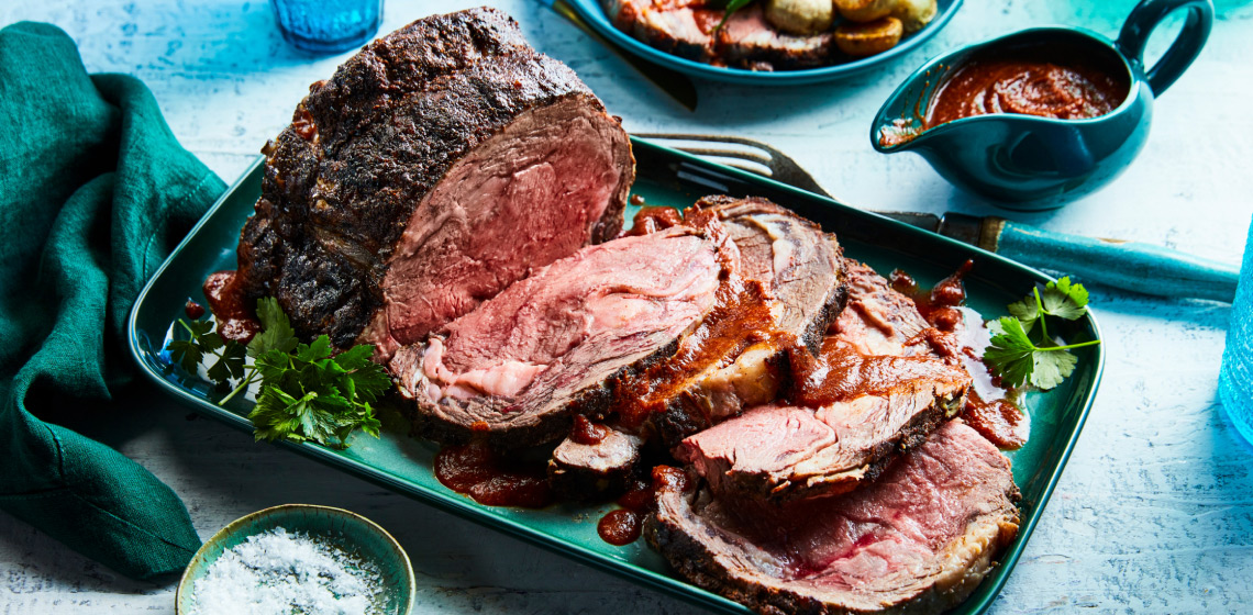 Rectangular aqua green serving platter with smoky Berbere spiced prime rib beef roast with slices cut from one end, sauce overtop and green herb garnish on the side