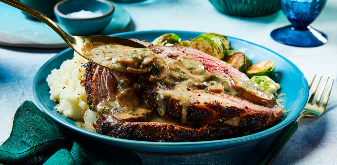 Round blue dinner plate with mashed potatoes, Brussels sprouts and Mushroom-Crust Prime Rib with Stroganoff Sauce being spooned overtop
