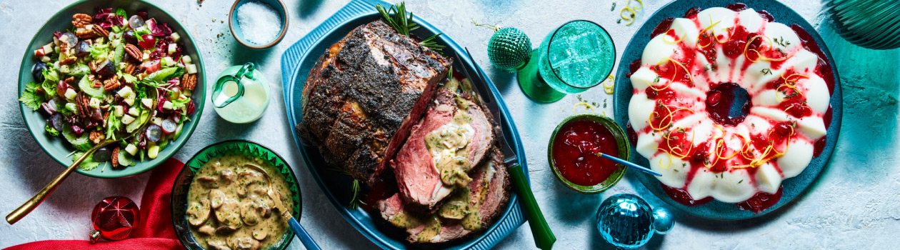 light blue surface with three serving platters showing a whole meal: roast beef, mushroom stroganoff sauce, waldorf-style salad and a luxurious cranberry-lemon blancmange pudding in bundt pan shape