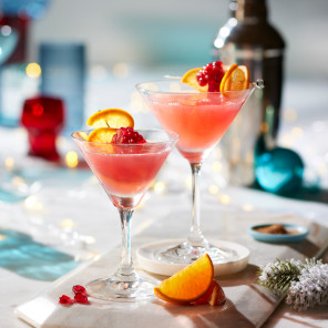 Easy holiday cocktails in two martini glasses with an orange and pomegranate garnish on a white marble surface with holiday decorations and a martini shaker in the background.