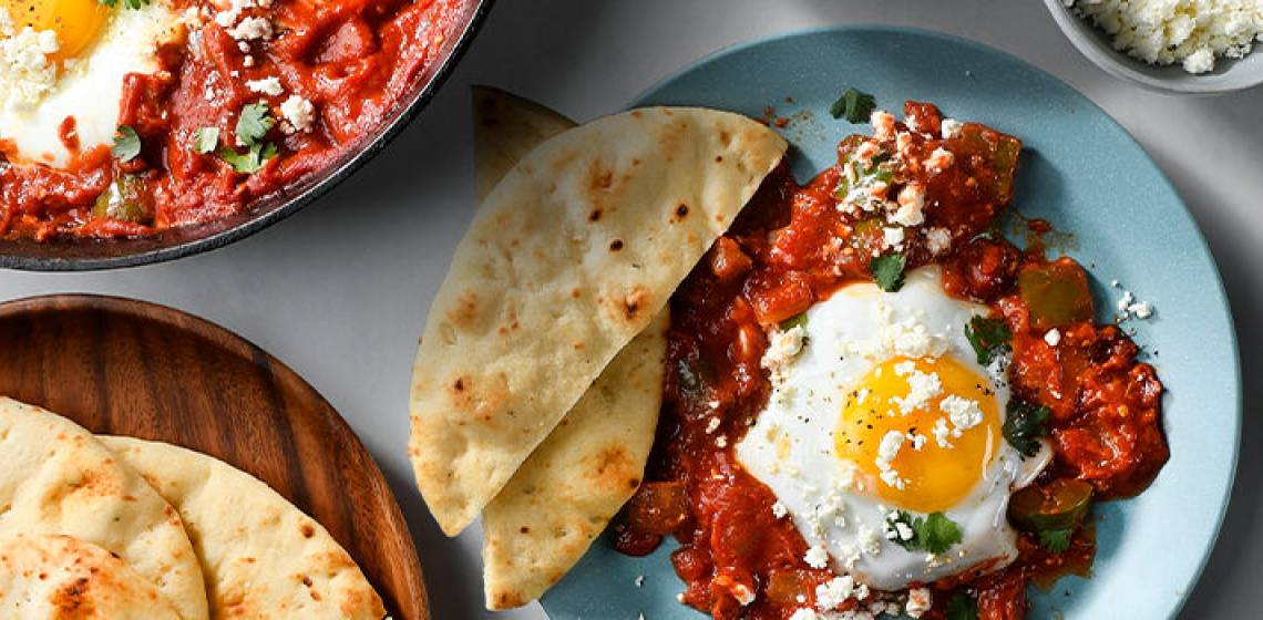 A light blue plate with one serving of shakshuka next to a cast-iron pan full of the dish on a white countertop.