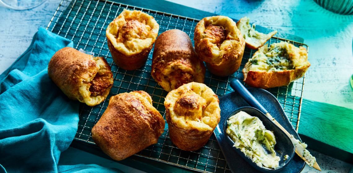 Aqua blue cutting board with metal cooling rack overtop, showing seven popovers and a side of herb butter