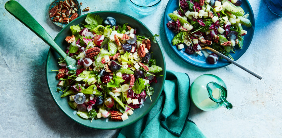 Large blue aqua-green bowl of waldorf-style salad with pecan garnish, alongside small blue side plate with single serving of salad and a side carafe of salad dressing.