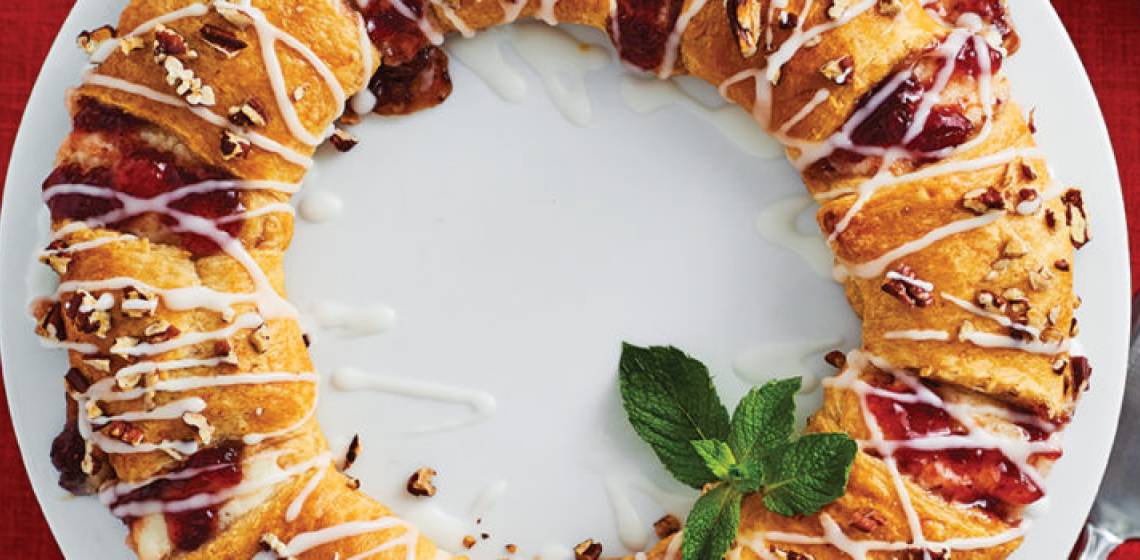 Round wreath-shaped strawberry jam, cream cheese, and lemon crescent-roll pastry on a large white platter on a red tablecloth-dressed table.