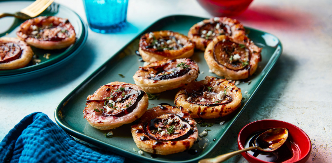 bite-sized rounds of baked puff pastry with fig, feta and balsamic glaze on a rectangular aqua serving platter with a blue napkin to the side