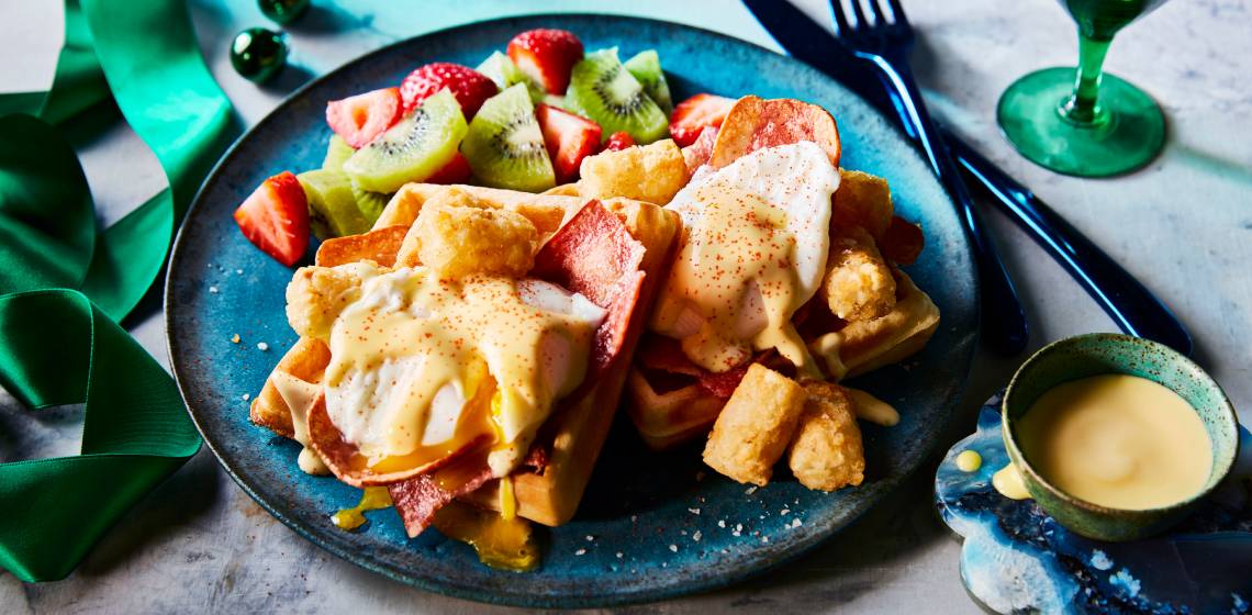 Belgian waffles served with a poached egg, turkey bacon-style, and potato tater nuggets, topped with hollandaise sauce on a dark blue plate next to a cup of coffee.