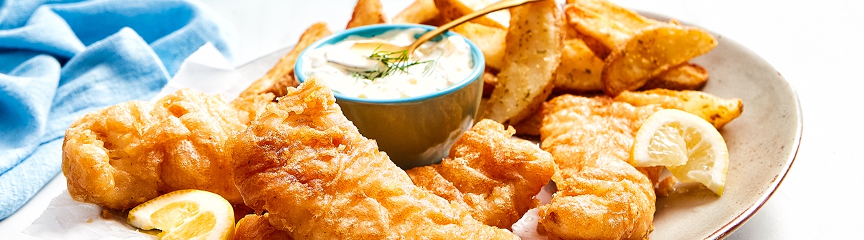 cream coloured oval platter with beer battered fish, French fries, lemon wedges and tartar sauce in a side bowl.