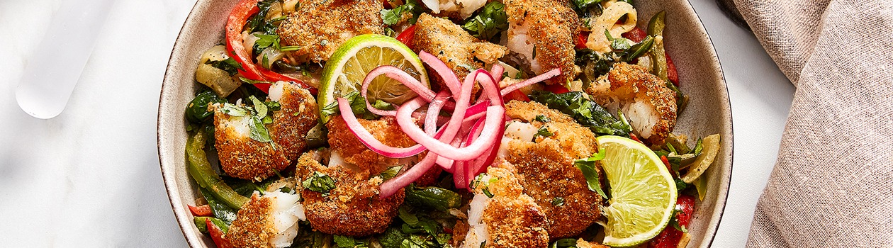 large shallow serving bowl of spinach bhorta with crispy cod nuggets, red peppers, lime wedges and pickled red onions on top.