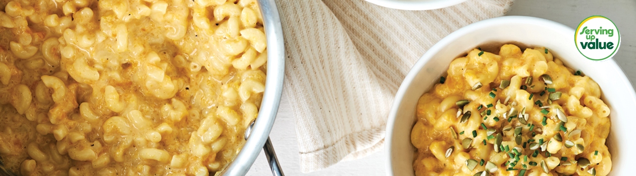 Stainless steel pot of butternut squash macaroni and cheese with two side bowls filled with pasta and a pistachio garnish.