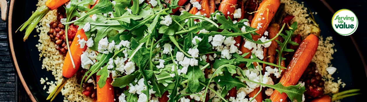 Large oval handled serving dish with couscous topped with lentils, roasted carrots, feta and fresh arugula.