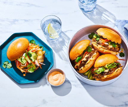 serving tray of three burgers, plus plated burger to the side, all stacked with a prime rib beef burger patty, pickled vegetables, cheese, and sriracha aioli on a brioche bun.