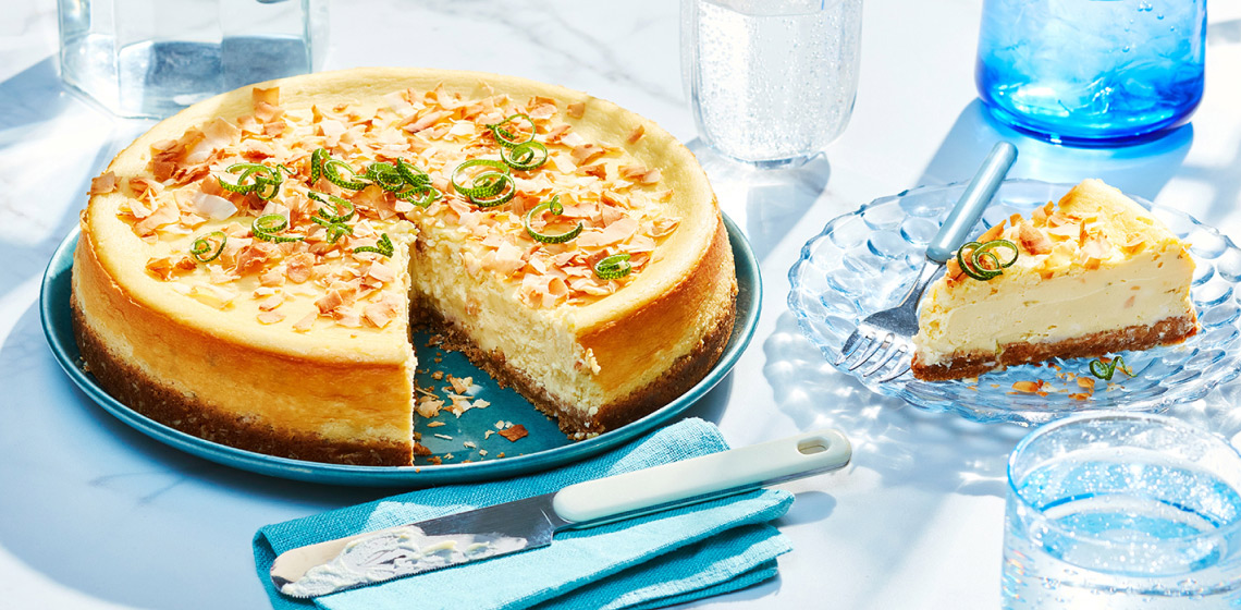 Full lime coconut cheesecake with lime peel garnish on serving plate with one slice removed and on plate to the right side.