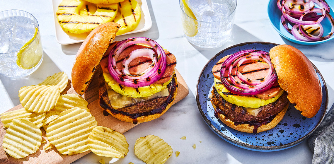 Two plates, each with Hawaiian-style burger topped with grilled pineapple round and side bowl of ruffled plain potato chips