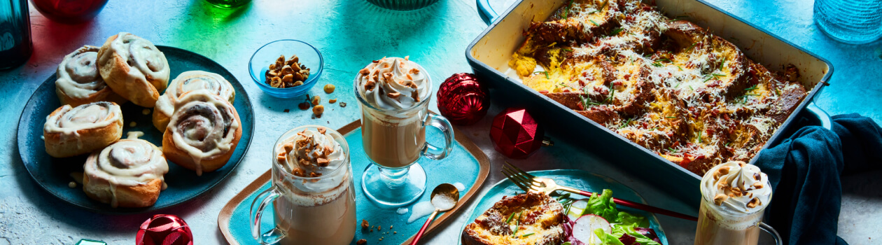 Light blue table surface with a serving plate of glazed cinnamon buns, eggnog french toast, two whip cream topped lattes, and a casserole dish of strata