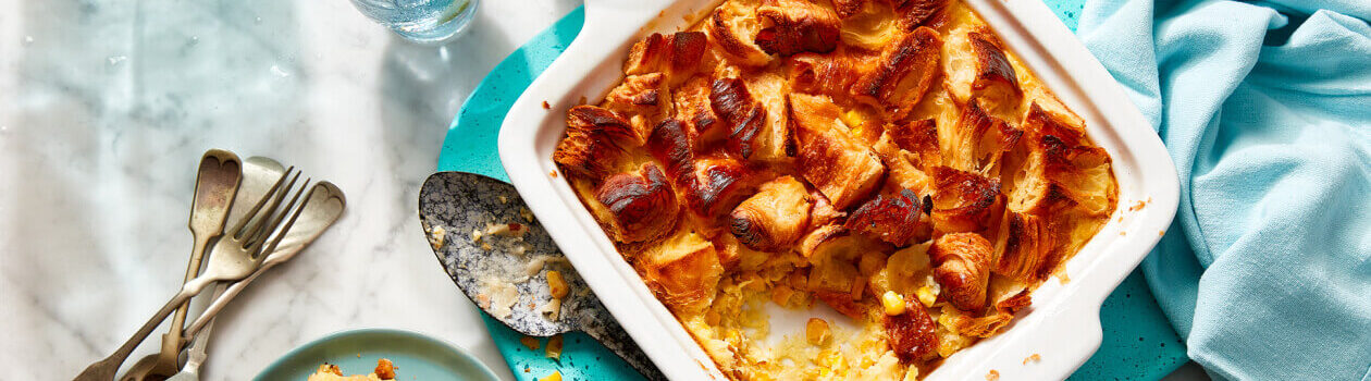 Croissant-topped corn pudding in a white baking dish with a serving next to it on a blue plate.