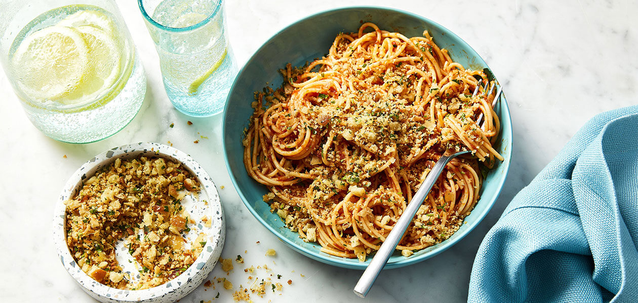 Italian-style Pangrattato seasoned breadcrumbs sprinkled over a simple tomato sauced pasta in a blue bowl with a smaller bowl full of just the breadcrumbs.