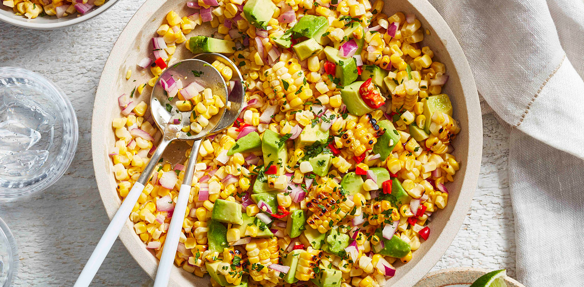 Overhead shot of Avocado & Corn Salad in a white serving bowl with two glasses of water and a small plate of cut limes next to it.