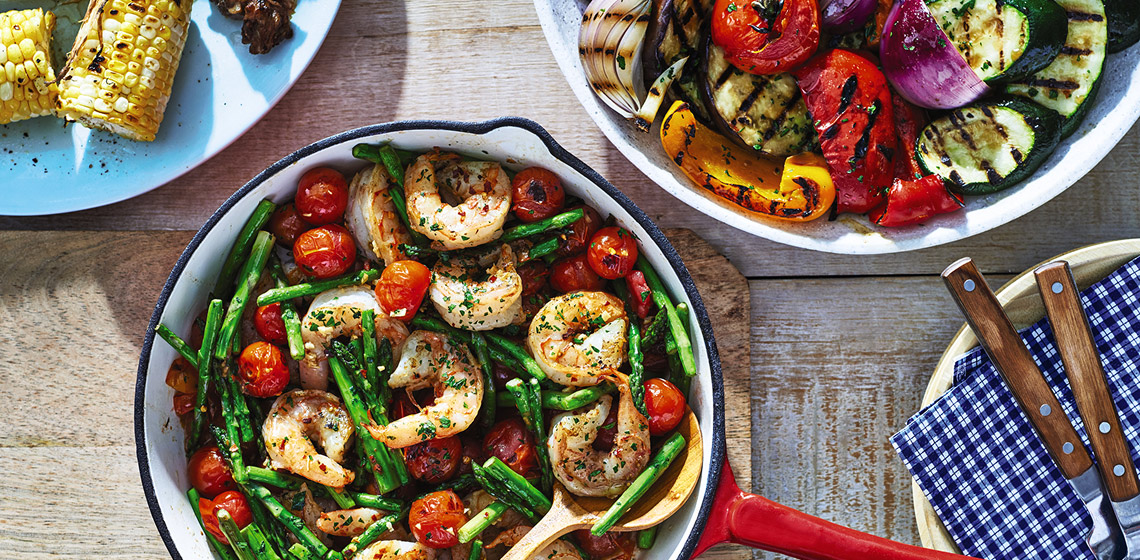 A red pot with lots of shrimp, asparagus, and cherry tomatoes with a wooden serving spoon.