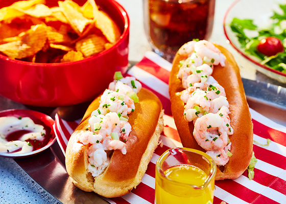 Two shrimp rolls on a white and red napkin with other bowls filled with salad and chips.