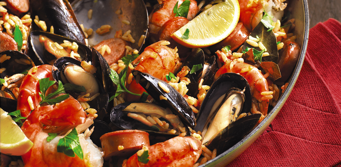 Light green pan filled with Barbecued Spanish Rice with Sausage & Seafood with a large serving spoon in it and a red towel folded underneath it.