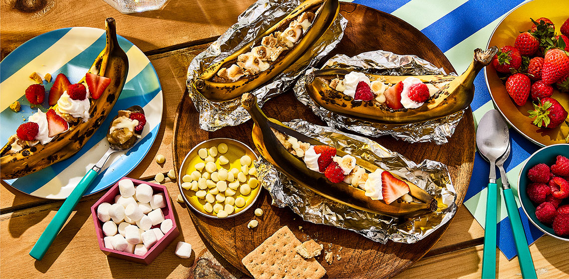 Berry shortcake banana boats on picnic table with plate of strawberries and raspberries to the side.