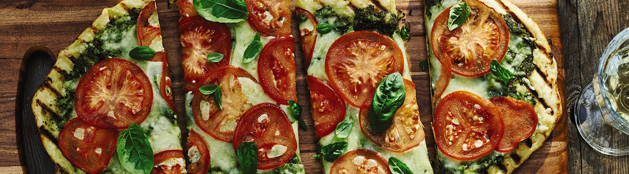 A cut up grilled tomato pizza on a wooden board with a pizza cutter also on the board and some filled glasses around it.
