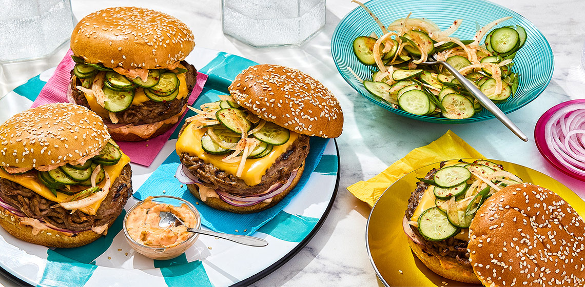 Three big Korean-style burgers and kimchi mayo on a blue and white striped plate, a Korean-style burger on a yellow plate and Korean-style pickled cucumber salad in a blue serving bowl on a marbled surface.