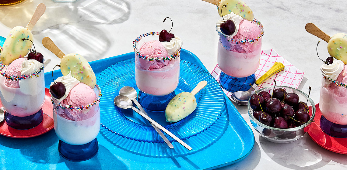 Five cups of the Birthday Cake Mini Bar Float with cherries in a bowl off to the side, spoons, a birthday cake mini bar, sprinkles, and various plates.