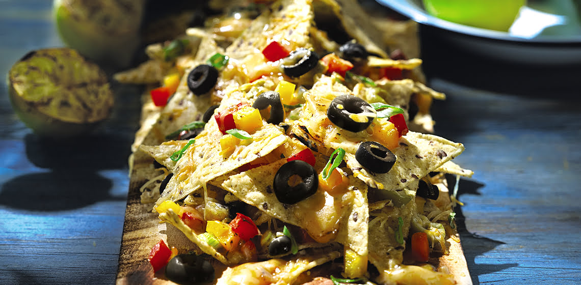 Nachos topped with cheese, olives, tomatoes, and peppers, served on a cedar plank