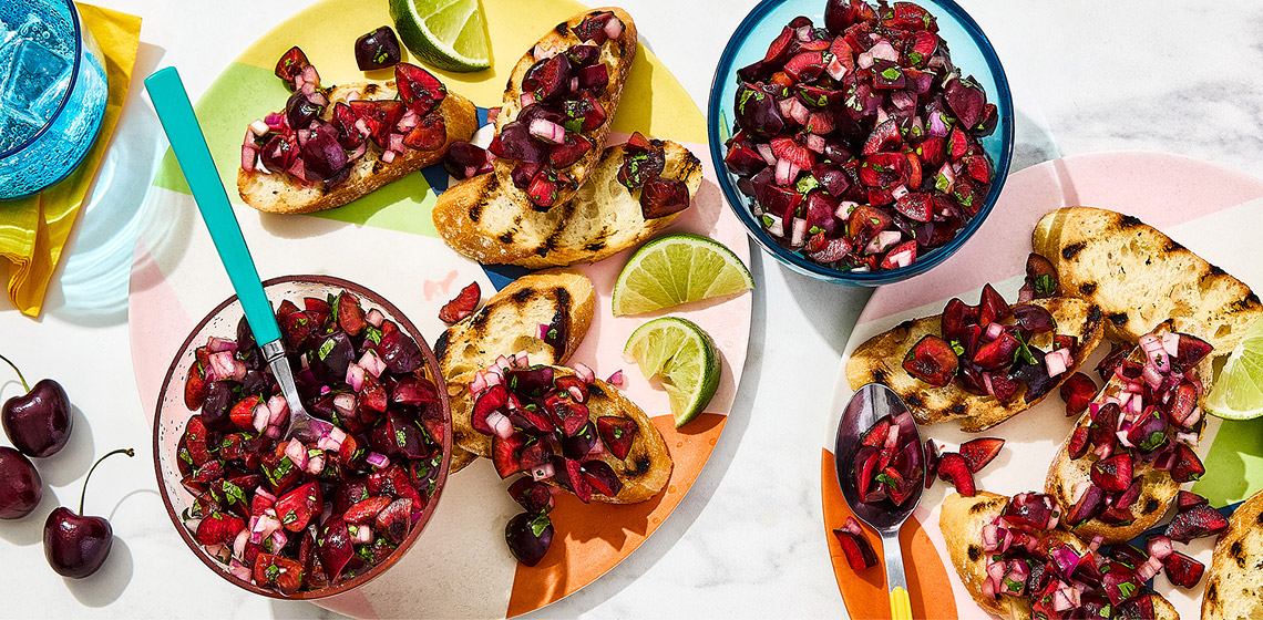 Two plates with crostinis topped with the cherry salsa and sliced limes, with bowls of the salsa and cherries on and around the plates.