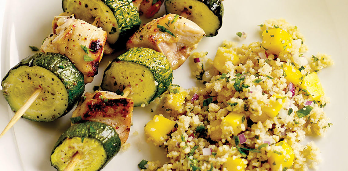 Two chicken and zucchini kabobs with tropical couscous on the side on a white plate.