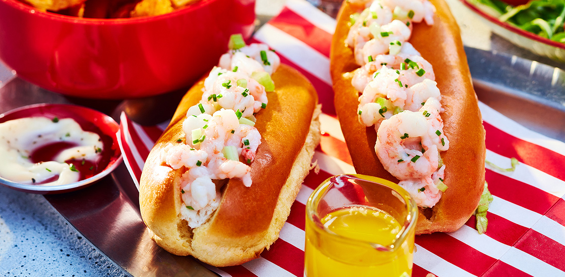 Two shrimp rolls on a white and red napkin with other bowls filled with salad and chips.