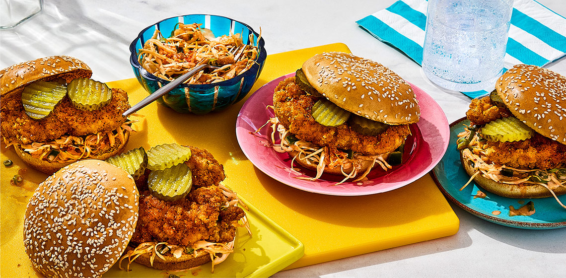 Chopped Korean fried chicken sandwiches topped with pickles, slaw, with a side of slaw in a small bowl to the side.
