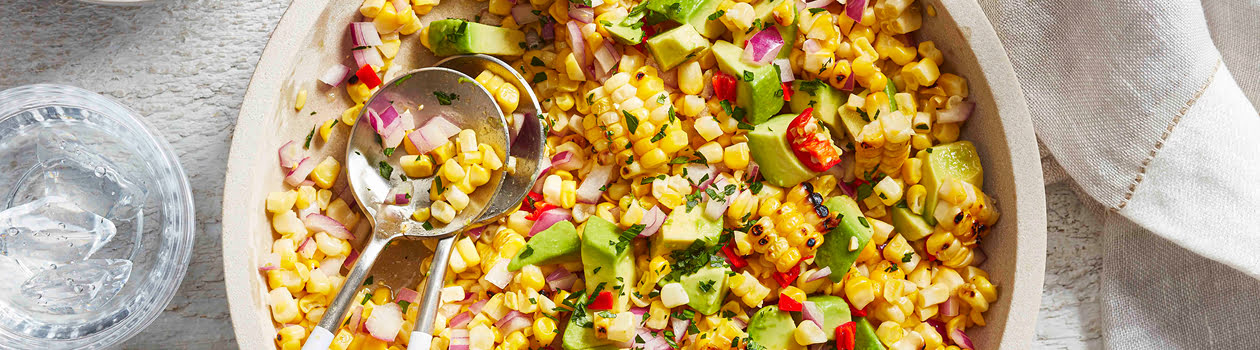 Overhead shot of Avocado & Corn Salad in a white serving bowl with two glasses of water and a small plate of cut limes next to it.