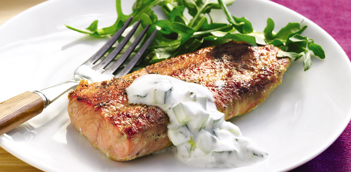 Plate of salmon with cucumber sauce, some arugula and a fork with more salmon and sauce in the background on a tray.