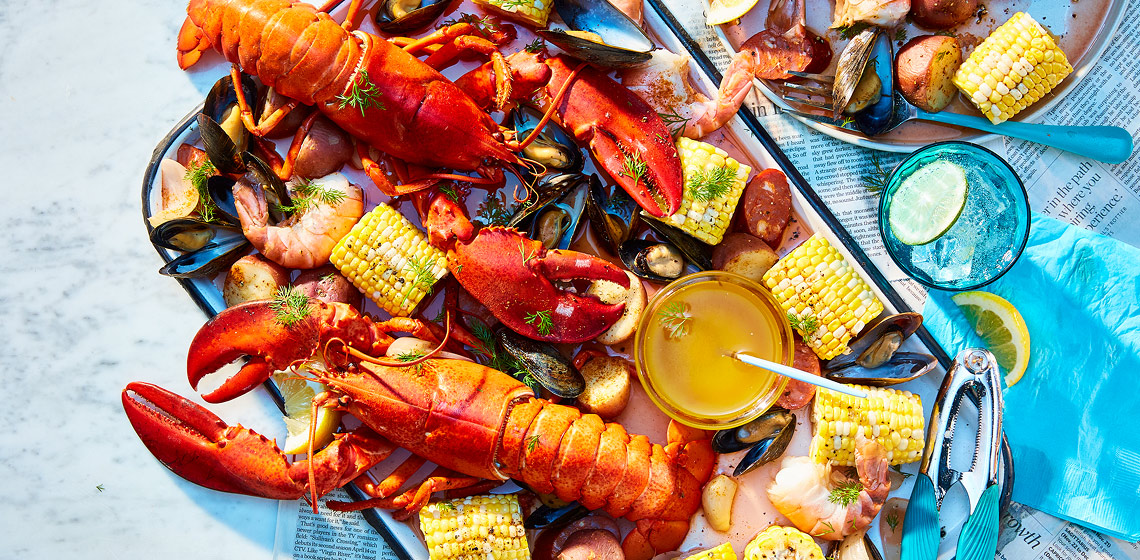 Large platter of a lobster boil with lobster, corn, shrimp, mussels, potatoes, sausage, and a bowl of sauce with a pair of small tongs.